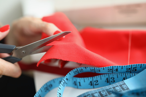 Female hands with manicure cut red fabric with scissors close-up. There is a cloth and a measuring tape on the table