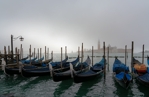 View on canal in Venice with anchored boats along the pier by residential buildings, one boat driving on the water surface, foggy winter day