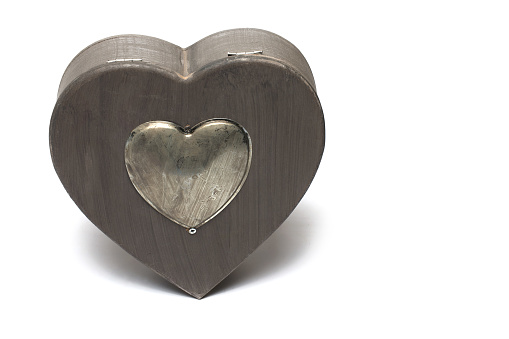 Close up of wooden box in the shape of heart with closed lid on which metal heart is attached on white background