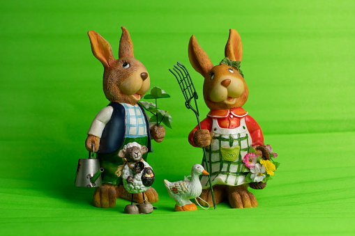 Group of ceramic figures, two rabbits with big bunny ears and gardening stuff, little sheep with basket of Easter eggs and small goose, standing together, green isolated background, front view.