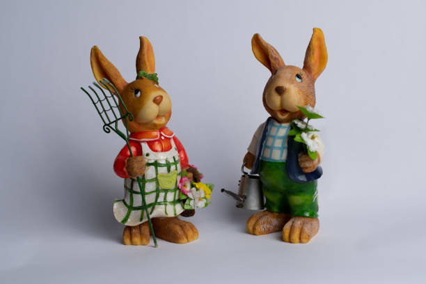 istock Ceramic brown Easter rabbits looking up, with spring flowers, rake and watering can in their hands, isolated on white background, front view. 1385720701