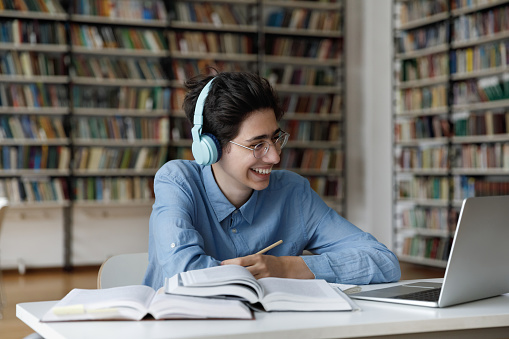 Happy young male Jewish student in eyeglasses wearing headphones, watching interesting educational lecture or webinar, studying on online courses, writing notes improving knowledge, e-learning.