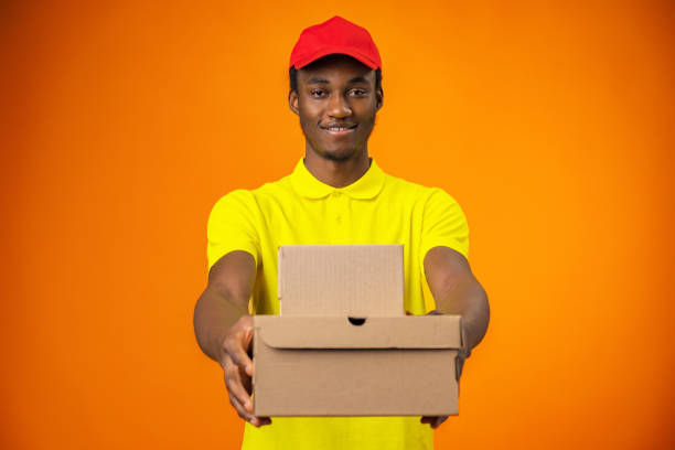 istock Smiling black male courier wearing uniform holding box in orange studio 1385724633