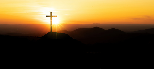 Religious grief landscape background banner panorama - Breathtaking view with black silhouette of mountains, hills, forest and cross / summit cross, in the evening during the sunset, with orange colored sky