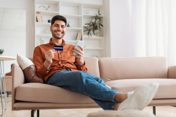 istock Smiling Arab guy using phone and credit card at home 1385727966
