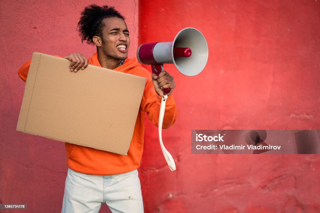 A male protestor using a megaphone and holding a blank placard An African-American man is holding a megaphone and a blank cardboard placard, wearing an orange sweatshirt against an orange background Activist Stock Photo