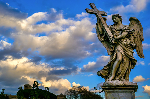 A beautiful and idyllic sunset sky behind a Bernini's angel along Ponte Sant'Angelo, in the historic and Baroque heart of Rome. This bridge was built over the River Tiber at the behest of the emperor Hadrian in 134 AD to connect the heart of the Ancient Rome with his mausoleum, currently Castel Sant'Angelo. A few centuries later, under the pontificate of Pope Clement IX, the architect and sculptor Giovanni Lorenzo Bernini was commissioned to create the current balustrade decorated with ten statues of the Angels of the Passion of Christ. According to the Christian tradition, each Angel holds one of the relics of the Passion of Christ, such as the cross, the veil of Veronica, the nails, the spear, the pillar of the scourging, etc. Designed by Bernini, the statues were made under his direction in 1669 by the pupils present in his school of sculpture. In 1980 the historic center of Rome was declared a World Heritage Site by Unesco. Super wide angle image in high definition format.