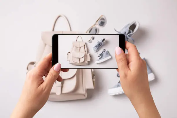 Woman taking photo of white leather backpack, pastel blue sneakers and sunglasses with smartphone. Blogger, influencer or stylist capturing spring fashion accessories for social media.