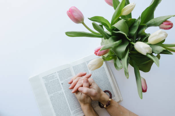 Hands folded in prayer, with chotki prayer bracelet on one wrist, on open bible with vase of pink and white tulips shot from above on a white background with copy space