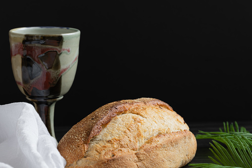 Clay chalice with loaf of artisan bread with palm leaves and white linen on a black background with copy space