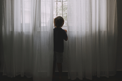 The boy looks out the window, hung with light curtains.