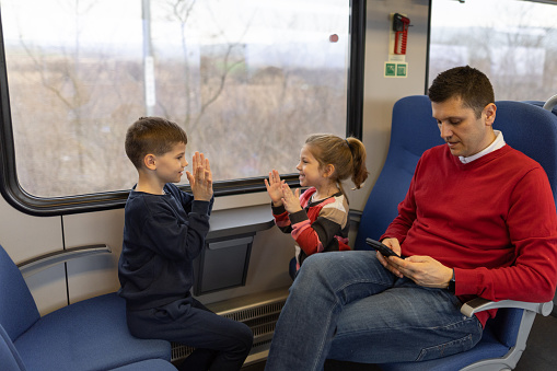 Cute little girl and her older brother are having fun playing together while riding on a train, their father is with them.