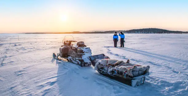 Parked snowmobile with trailer sled full of supplies covered with reindeer fur on the frozen Lake Inari in Finland, Lapland. Winter scene with the back of two unrecognizable people enjoying the beautiful sunrise. Panoramic view.