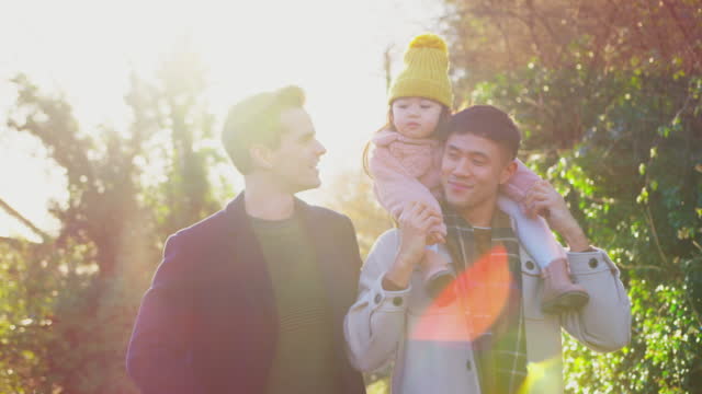 Family with two dads on walk in winter countryside carrying daughter on shoulders  against flaring sun - shot in slow motion