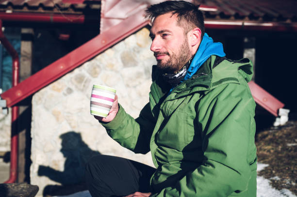 Attractive young man holding cup of hot beverage outdoors in cold but sunny winter morning. Trying to warm-up body with tea or coffee.