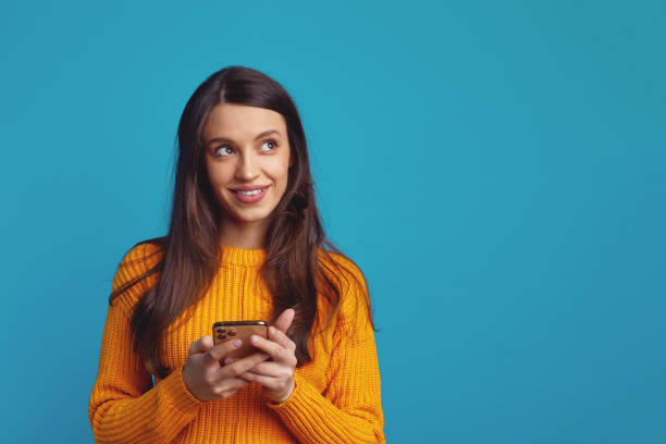 Cute girl holds mobile phone has thoughtful expression, looks away against blue Dreamy young woman holds mobile phone has thoughtful expression thinks about received message looks away isolated over blue background mobile phone text messaging telephone women stock pictures, royalty-free photos & images