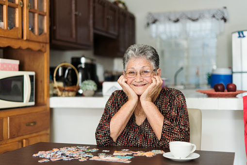 Happy senior woman making a puzzle, sitting at a table at home and looking at camera.