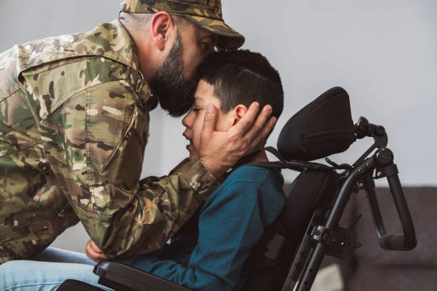 un padre soldado besa a su hijo discapacitado en una silla de ruedas antes de ir a la guerra - concepto de familia disfuncional - - personal militar español fotografías e imágenes de stock