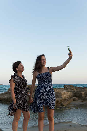 In a coastal area, at sunset, a young girl and her mother are taking a selfie on the seashore, next to some rocks.