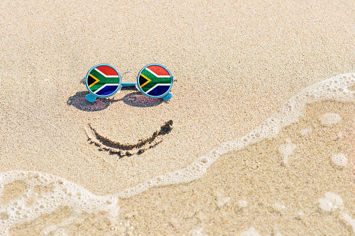 A painted smile on the sand and sunglasses with the flag of South Africa. The concept of a positive and successful holiday in the resort of South Africa.