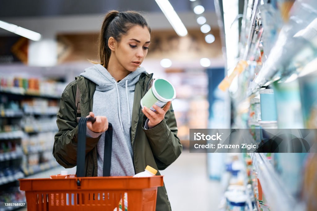 Young woman reading nutrition label while buying diary product in supermarket. Young woman buying diary product and reading food label in grocery store. Supermarket Stock Photo
