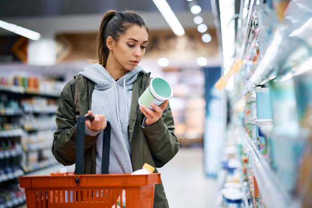 Young woman reading nutrition label while buying diary product in supermarket. Young woman buying diary product and reading food label in grocery store. refrigerated section supermarket photos stock pictures, royalty-free photos & images