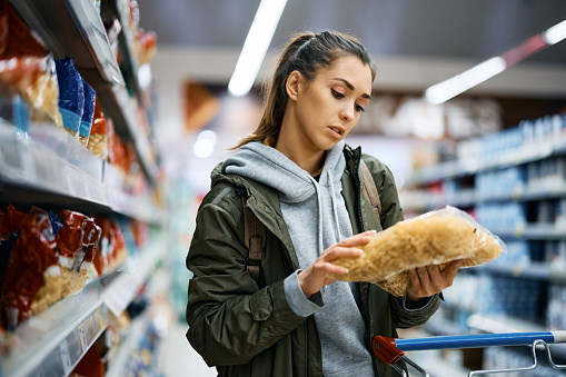 Young woman reading nutrition label while buying pasta at supermarket.
