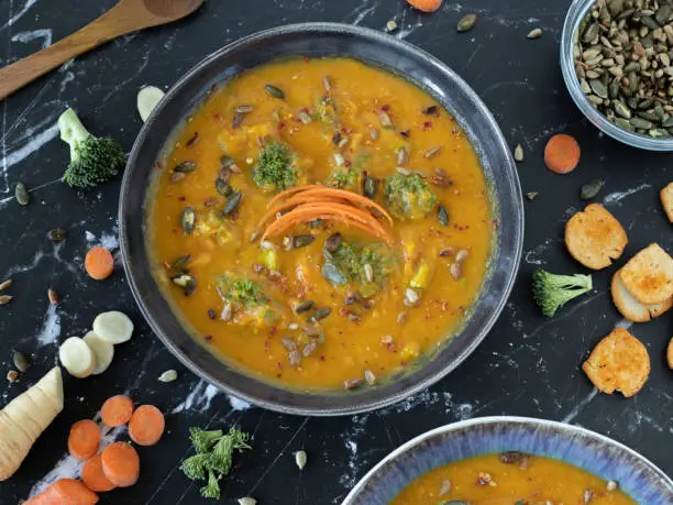 Orange vegan plant-based soup in a bowl with a wooden spoon and various chopped vegetables on a dark granite table. The concept of cooking and eating healthy food. Top view.