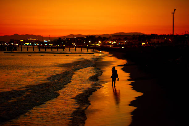 The lady is walking on the beach. She is covered by the crimson light of the setting sun.