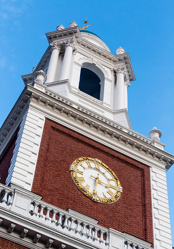 Cambridge, Massachusetts, USA - January 19, 2022: Close-up of Eliot House clock tower (c. 1931) against a blue sky. Gold numbers and hands on the clock face.