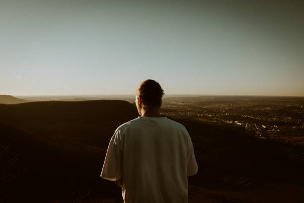 Young man standing alone on mountain top enjoying the view during twilight and the setting sun. Thinking and planning in a silent, relaxed moment. Shot from behind. Millennial Generation Outdoors Lifestyle Portrait.