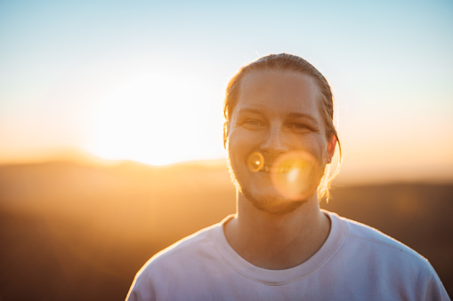Having Fun. Happy bright smiling young blonde man in white shirt looking outdoors towards the camera backlit from the sunset sun. Real People Millennial Generation Outdoors Lifestyle Portrait.