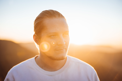 Confident slightly smiling young millennial generation blonde man in white shirt looking over towards his friends. Moody Sunset Light, Back Lit. Real People Millennial Generation Outdoors Lifestyle Portrait.