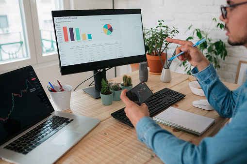 Close up of a successful Caucasian male freelancer casually working from the comfort of his home office, using a desktop computer, laptop and a smart phone to follow the current events related to stocks and cryptocurrency