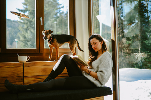 Young woman reading or studying in the mountain cabin with her pet dog resting next to her.