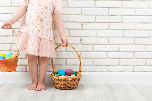Unrecognizable girl with colorful Easter eggs in two wicker baskets near white brick wall. Easter traditional egg hunt. Copy space