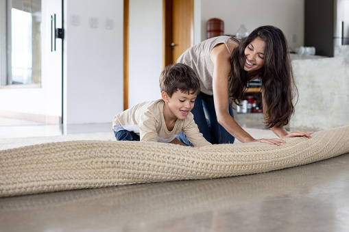 Happy Brazilian boy helping his mother unroll a carpet at home and smiling - teamwork concepts