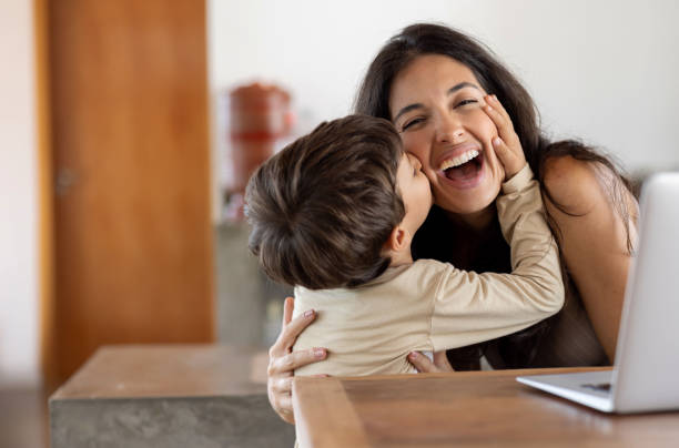 hijo amoroso dándole un beso a su madre mientras ella está trabajando en casa - hijos fotografías e imágenes de stock