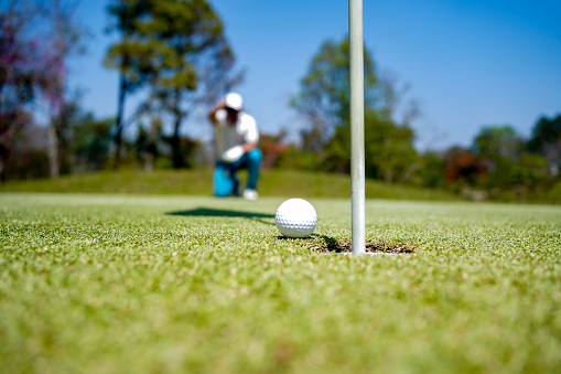 Select focus. Golfer asian man with friend playing aiming shot for putting ball on the hole with club on green course.  Hobby in holiday and vacations in sunny morning day on club golf. Lifestyle and Sport Concept