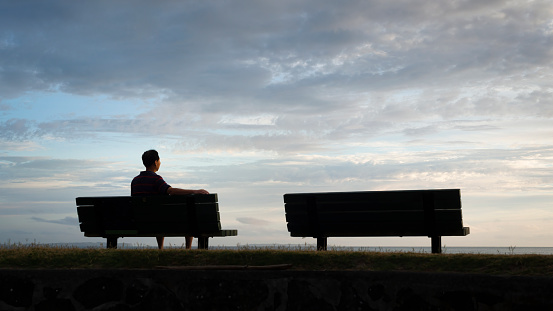 Man sitting on the bench facing the sea at dawn. Another empty bench by the side.