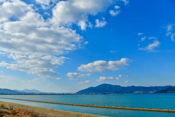 A serene lakeside scene featuring calm waters, a small settlement, and distant mountains partially obscured by low-hanging clouds. The foreground showcases a mix of farmland and scattered buildings, creating a harmonious blend of nature and human habitation. Warm hues of autumn foliage contrast against the tranquil blue tones of the lake.