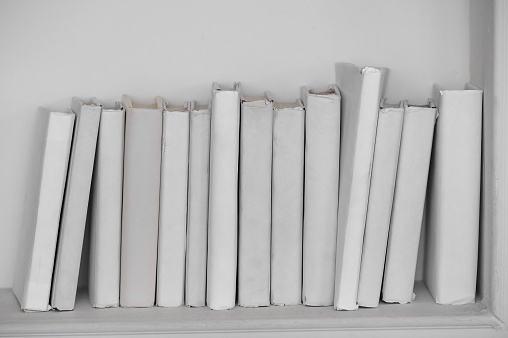 A row of all white books sit on a white shelf in front of an off white background.