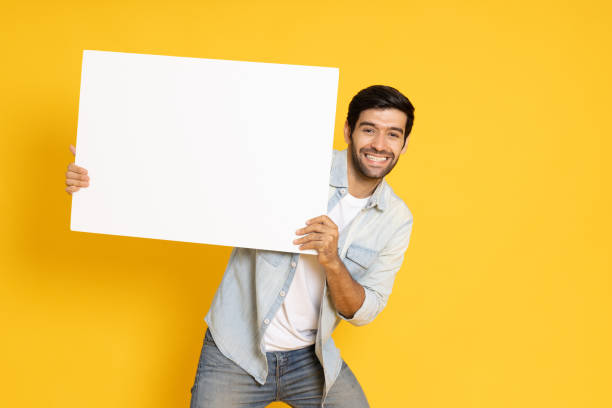 istock Young man is holding white blank banner or empty copy space advertisement board on yellow background, Looking at camera 1385911207
