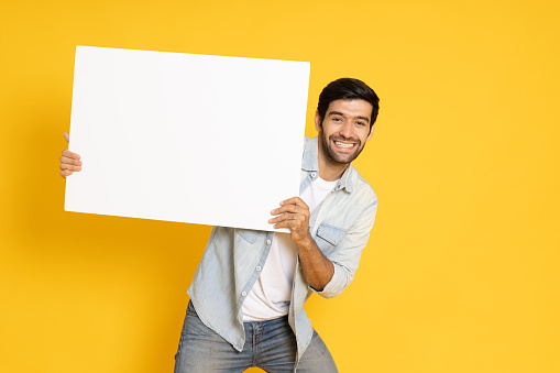 Young man is holding white blank banner or empty copy space advertisement board on yellow background, Looking at camera