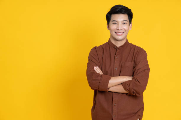 黄色いスタジオの孤立した背景で腕を組んで微笑む若いアジア人男性 - asia ストックフォトと画像