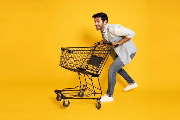 Photo of Full length portrait of young Caucasian man pushing an empty shopping cart or shopping trolley isolated on yellow background