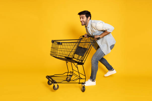 retrato completo de um jovem caucasiano empurrando um carrinho de compras vazio ou carrinho de compras isolado em fundo amarelo - running on empty - fotografias e filmes do acervo