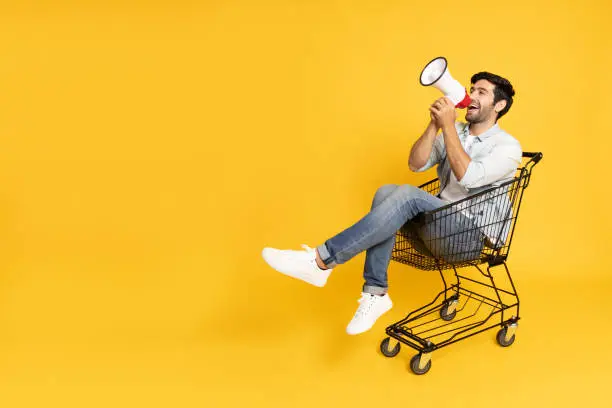 Young man sitting inside of shopping trolley and holding megaphone  isolated on yellow background, Announce concept