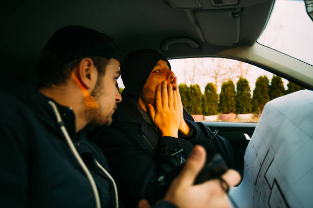istock Two armed bandits sitting in a car planning their next robbery, while counting on a stopwatch the time they have for the robbery, showing off their guns and pointing at the blueprint. 1385920281