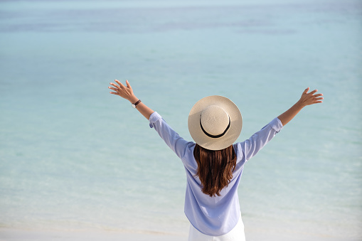 Rear view image of a woman with hat opening arms while walking on the white beach
